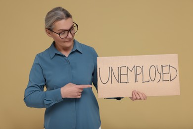 Senior woman pointing at cardboard sign with word Unemployed on beige background