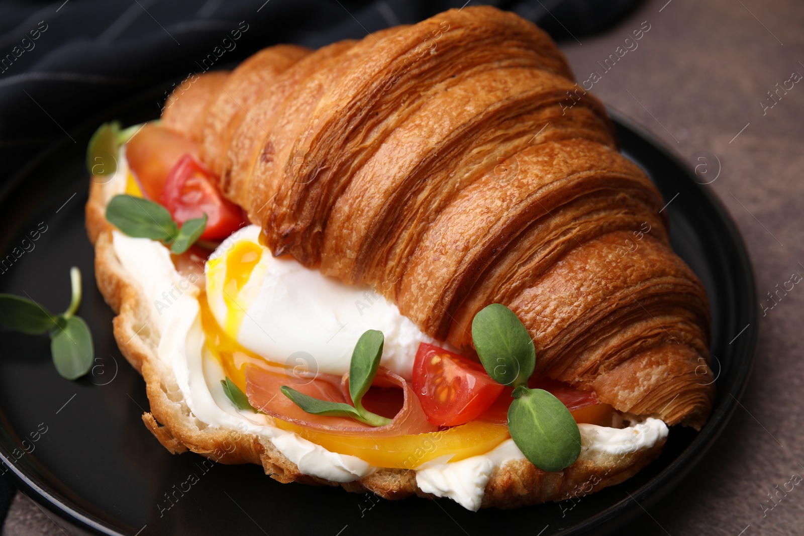 Photo of Tasty croissant with fried egg, tomato and microgreens on brown textured table, closeup