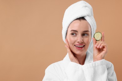 Photo of Beautiful woman in bathrobe with piece of cucumber on beige background, space for text