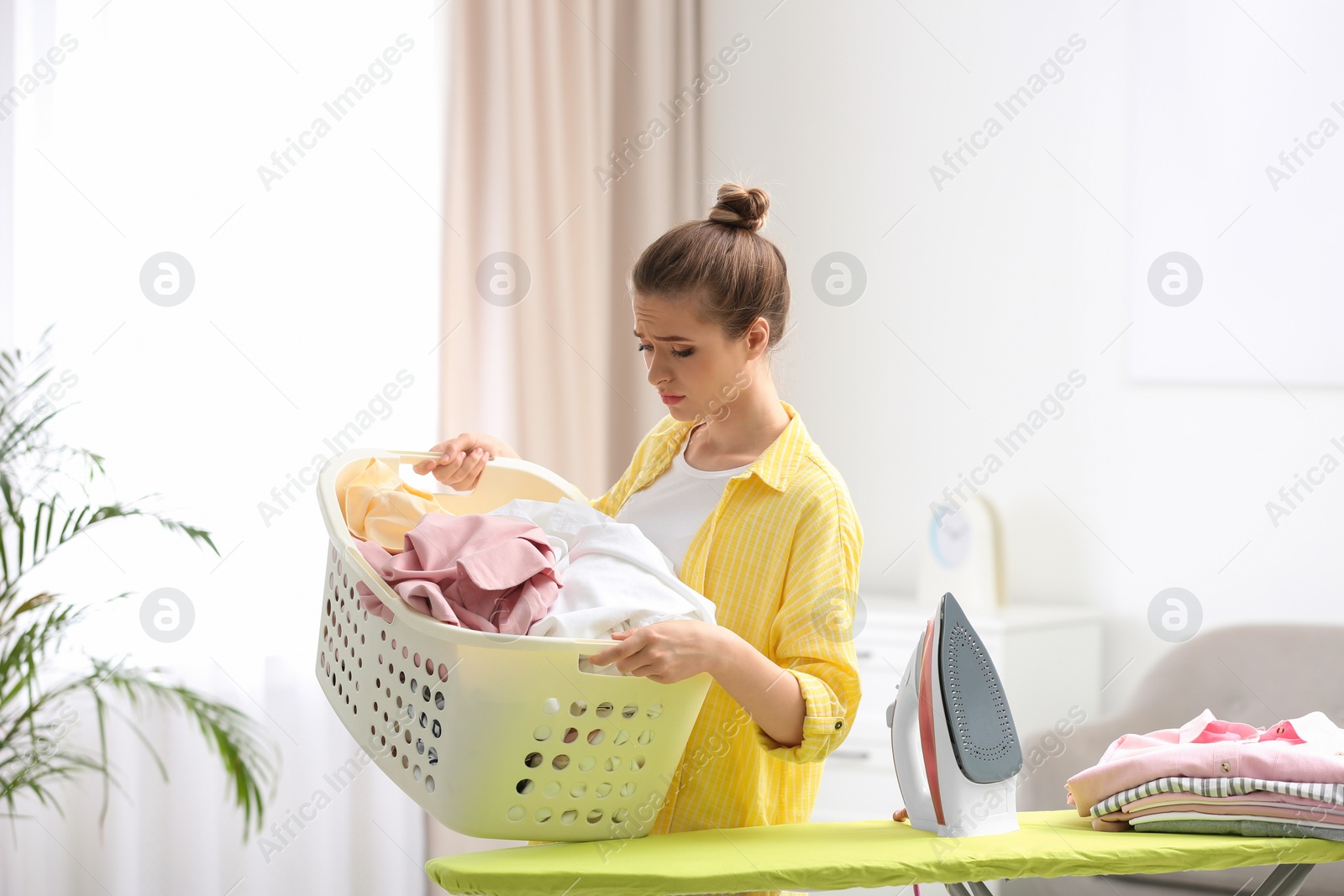 Photo of Young tired woman holding basket of clean laundry at ironing board indoors