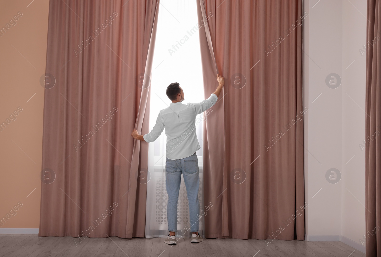 Photo of Man opening window curtains at home, back view