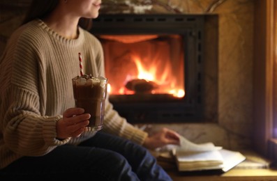 Woman with glass of hot cocoa and book near fireplace at home, closeup. Space for text