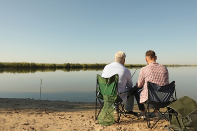 Father and adult son fishing together from riverside on sunny day