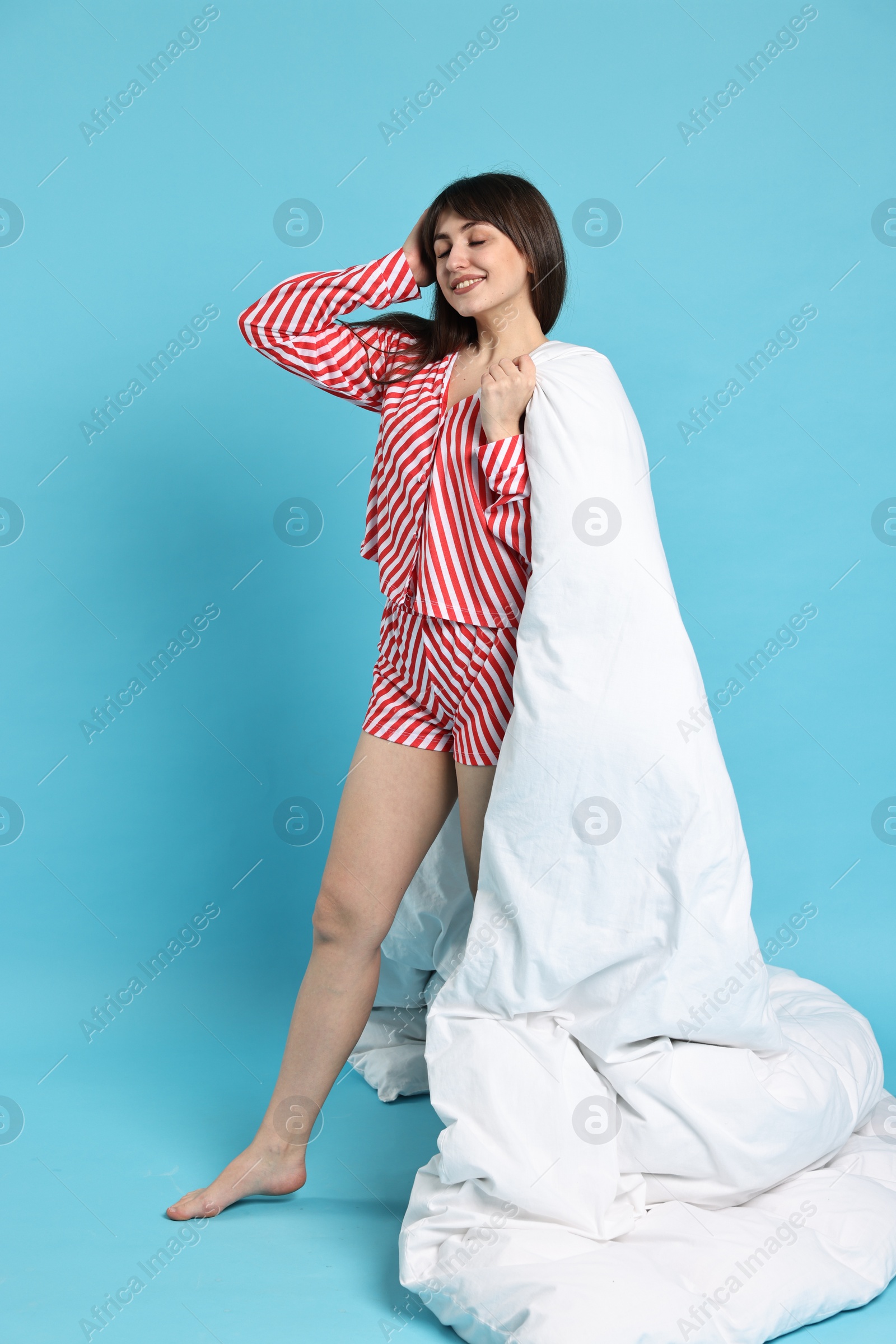 Photo of Happy woman wearing pyjama and holding blanket on light blue background