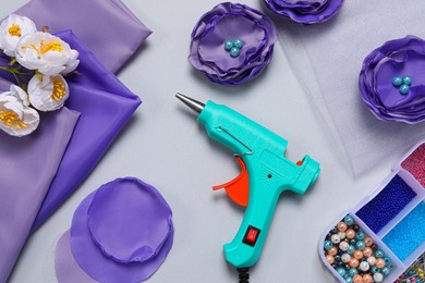 Photo of Hot glue gun, textile flowers and handicraft materials on grey background, flat lay
