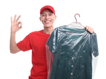 Photo of Dry-cleaning delivery. Happy courier holding dress in plastic bag and showing OK gesture on white background