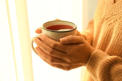 Photo of Woman holding elegant cup with tea near window indoors, closeup