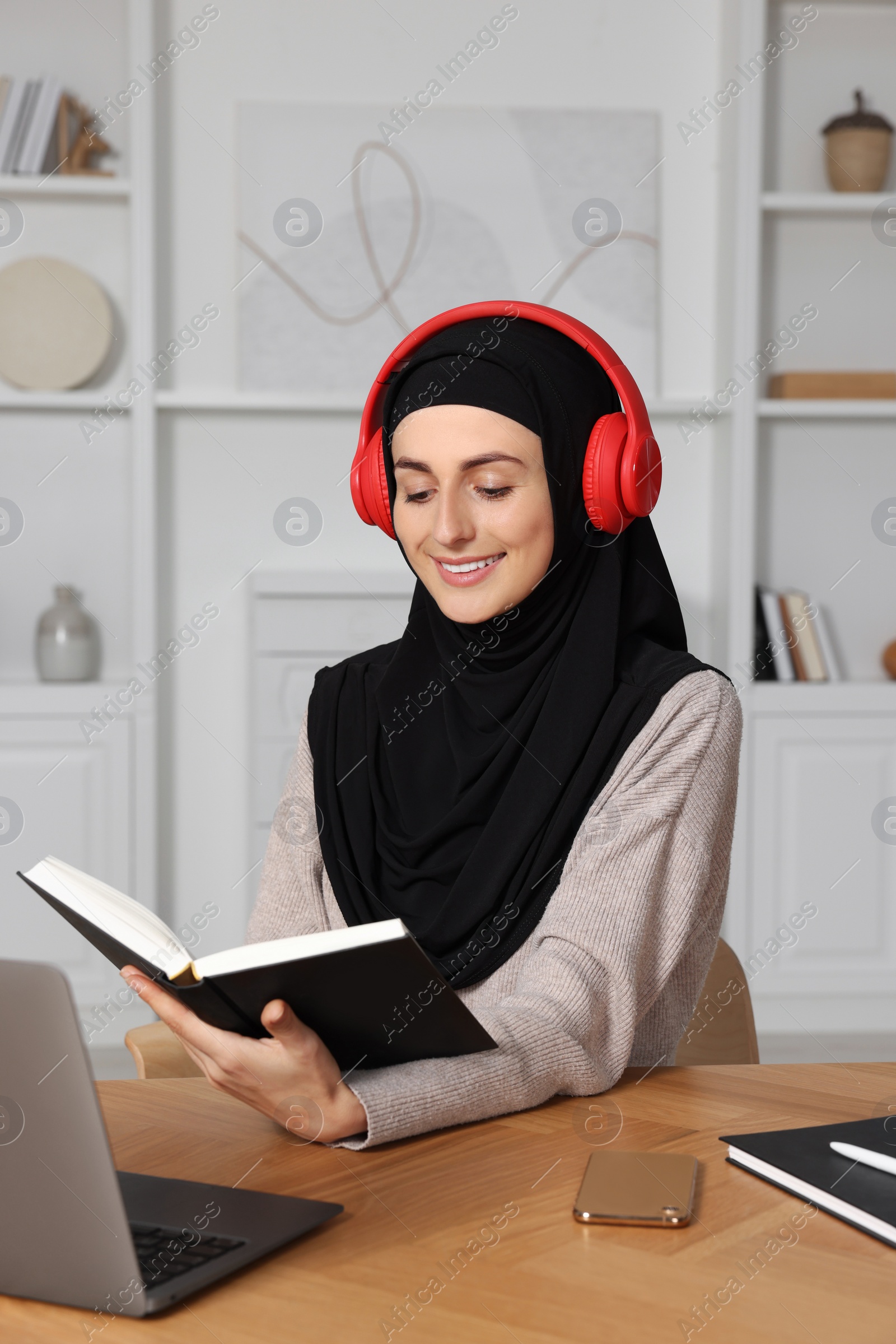 Photo of Muslim woman in headphones studying near laptop at wooden table in room