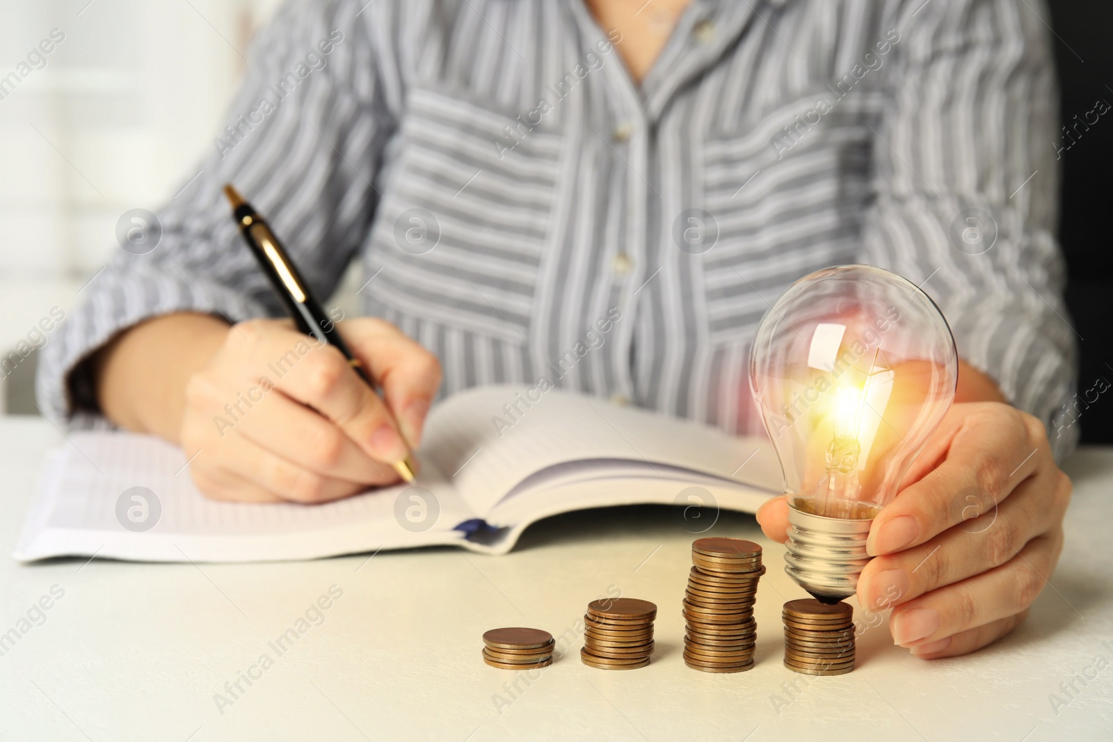 Photo of Woman with light bulb, notebook and coins at white table, closeup. Power saving