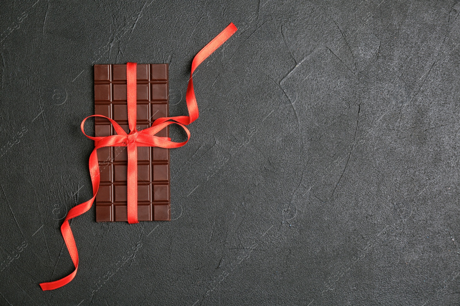 Photo of Tasty chocolate bar with red ribbon on dark background, top view