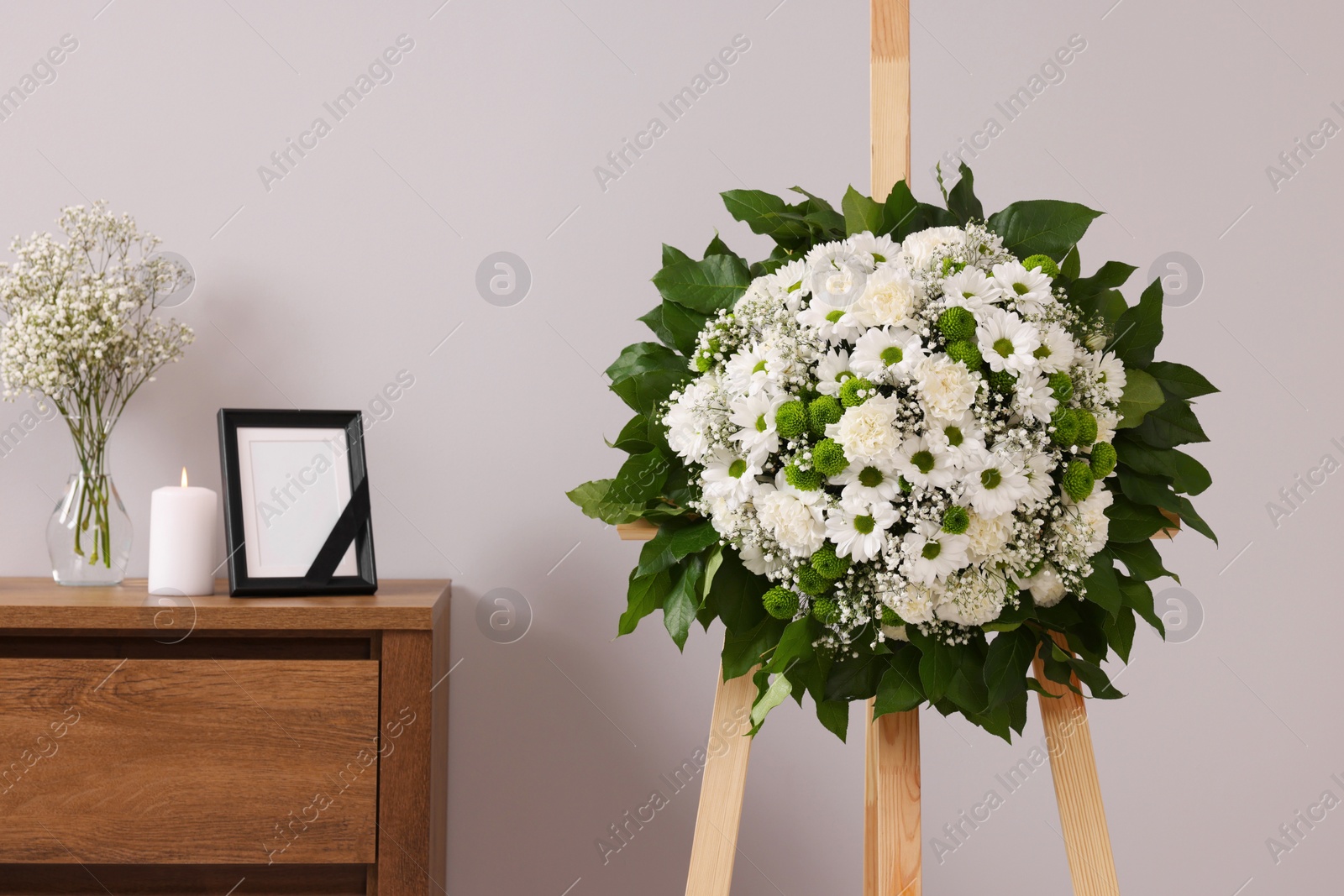 Photo of Wreath of flowers and frame with black ribbon on commode in room. Funeral attributes