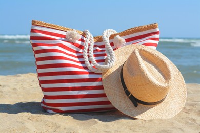 Stylish striped bag with straw hat on sandy beach near sea