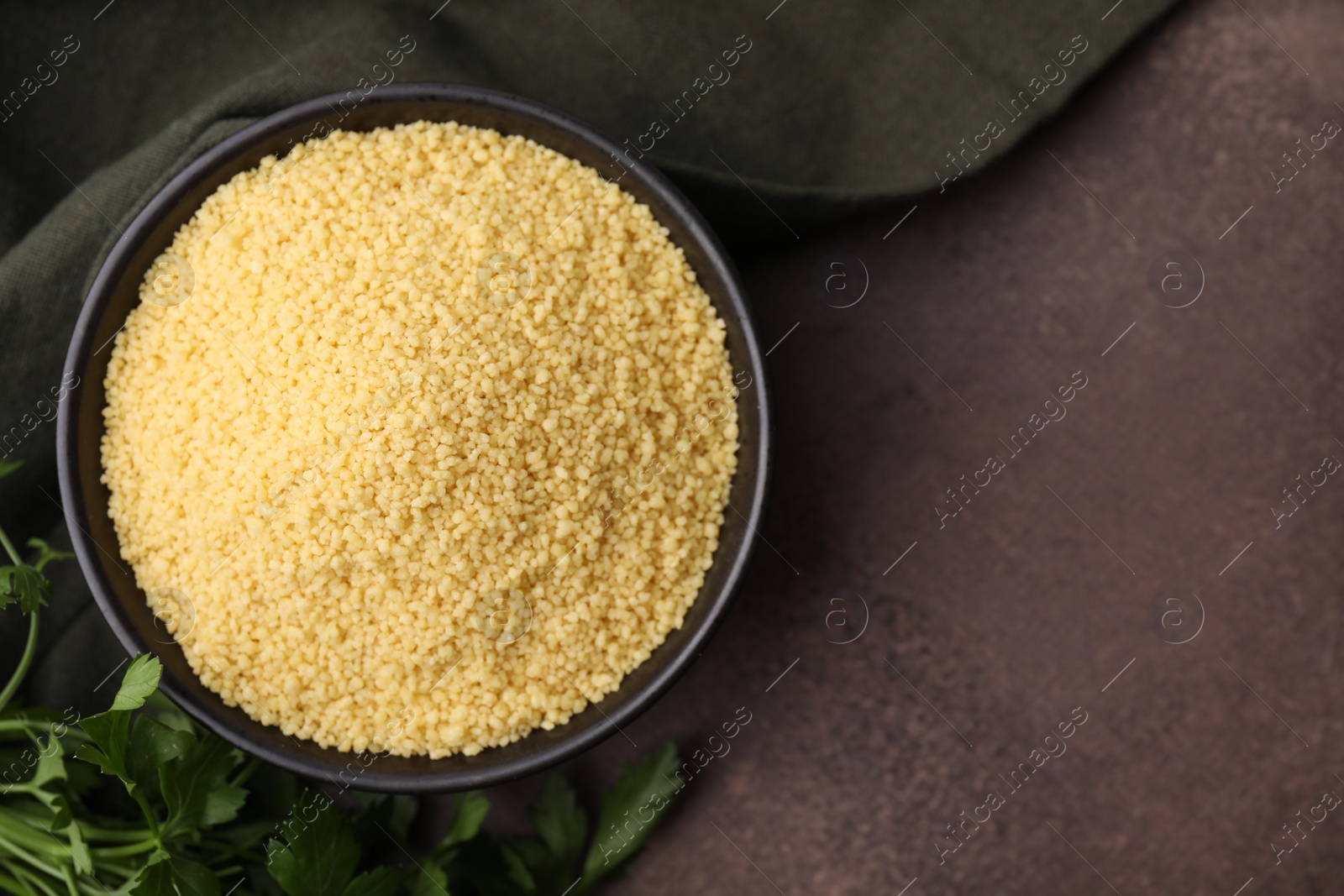 Photo of Raw couscous in bowl and parsley on brown table, top view. Space for text