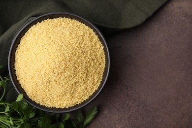 Raw couscous in bowl and parsley on brown table, top view. Space for text