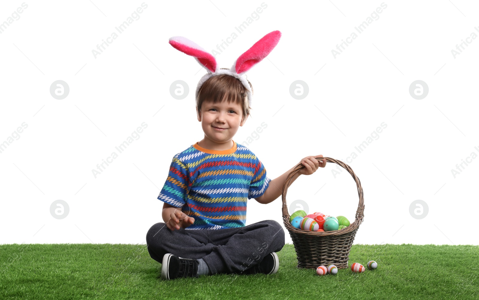 Photo of Cute little boy wearing bunny ears with basket full of dyed Easter eggs on green grass