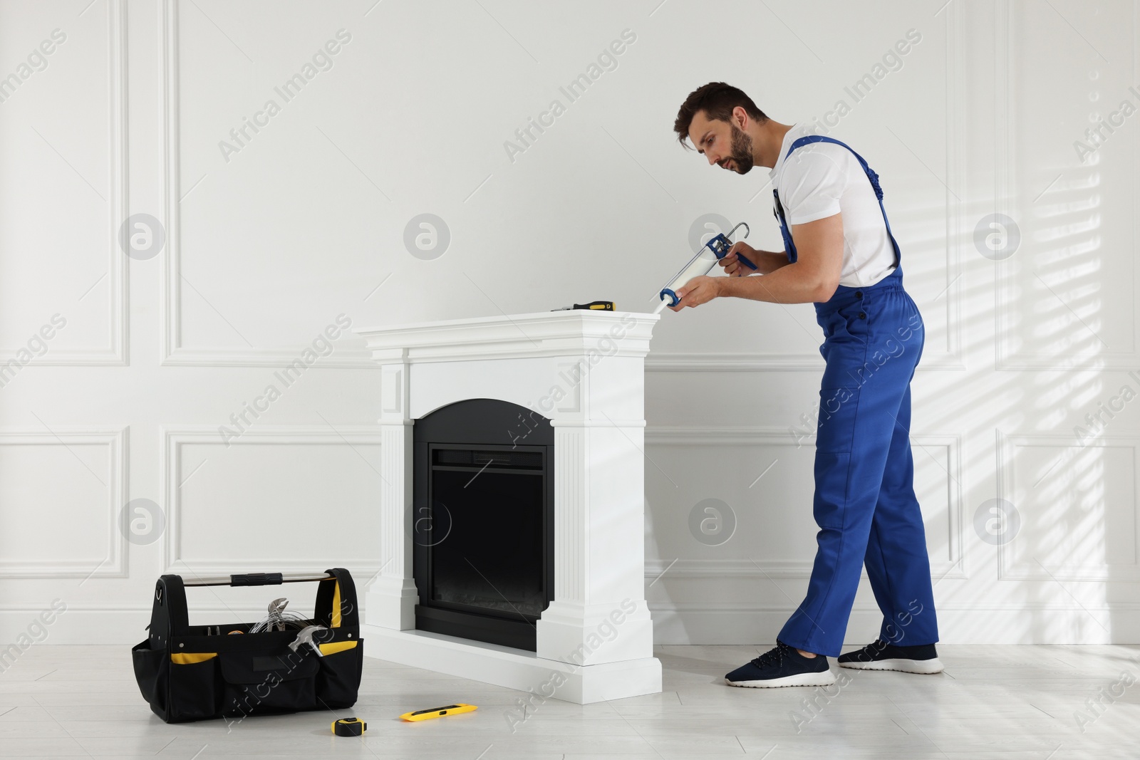 Photo of Professional technician sealing electric fireplace with caulk near white wall in room