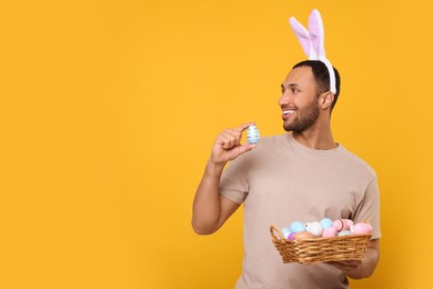 Happy African American man in bunny ears headband holding wicker tray with Easter eggs on orange background, space for text