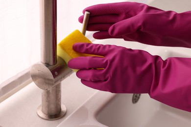 Photo of Woman in gloves cleaning faucet of kitchen sink with sponge, closeup