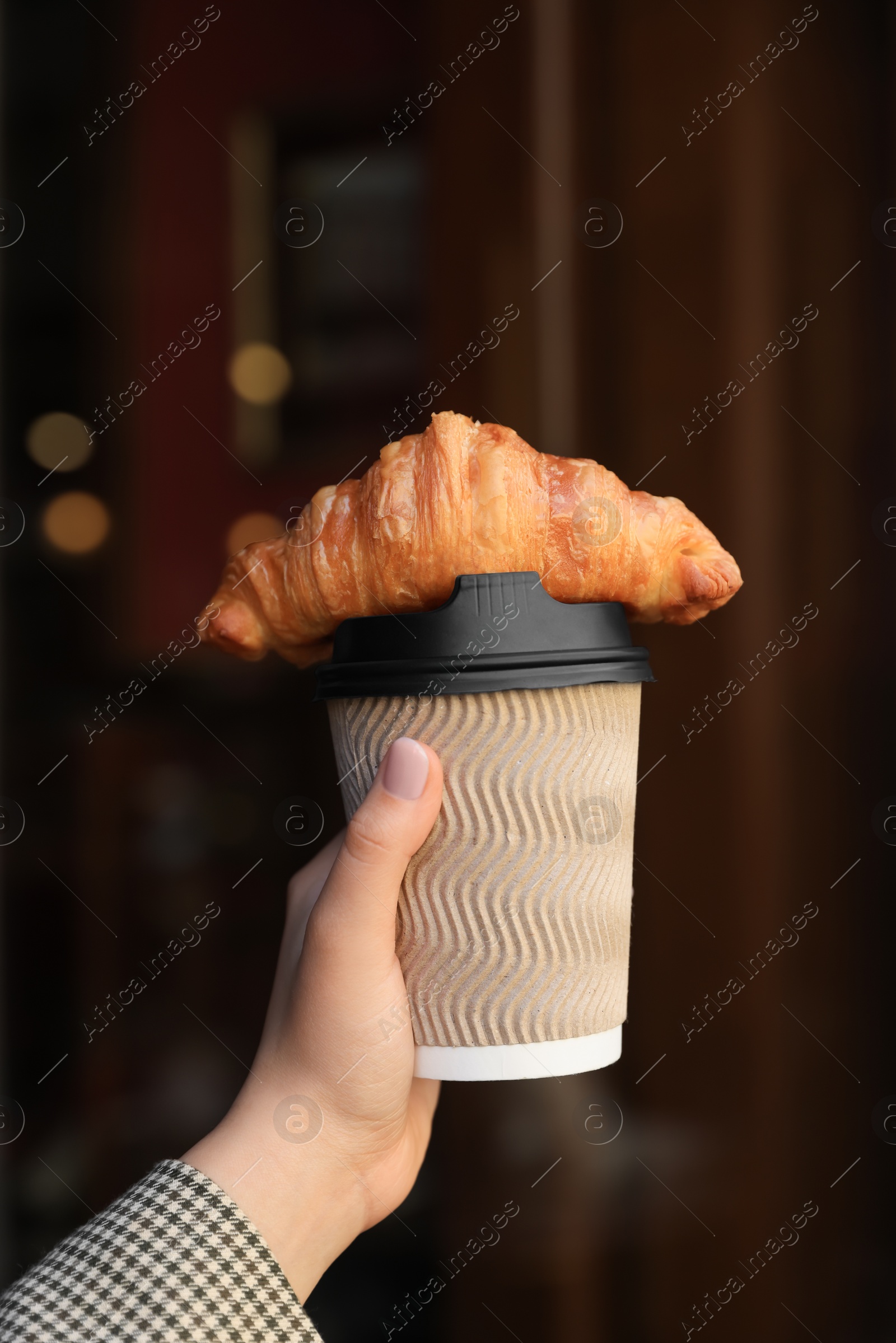 Photo of Woman holding tasty croissant and cup of coffee near wooden door outdoors, closeup
