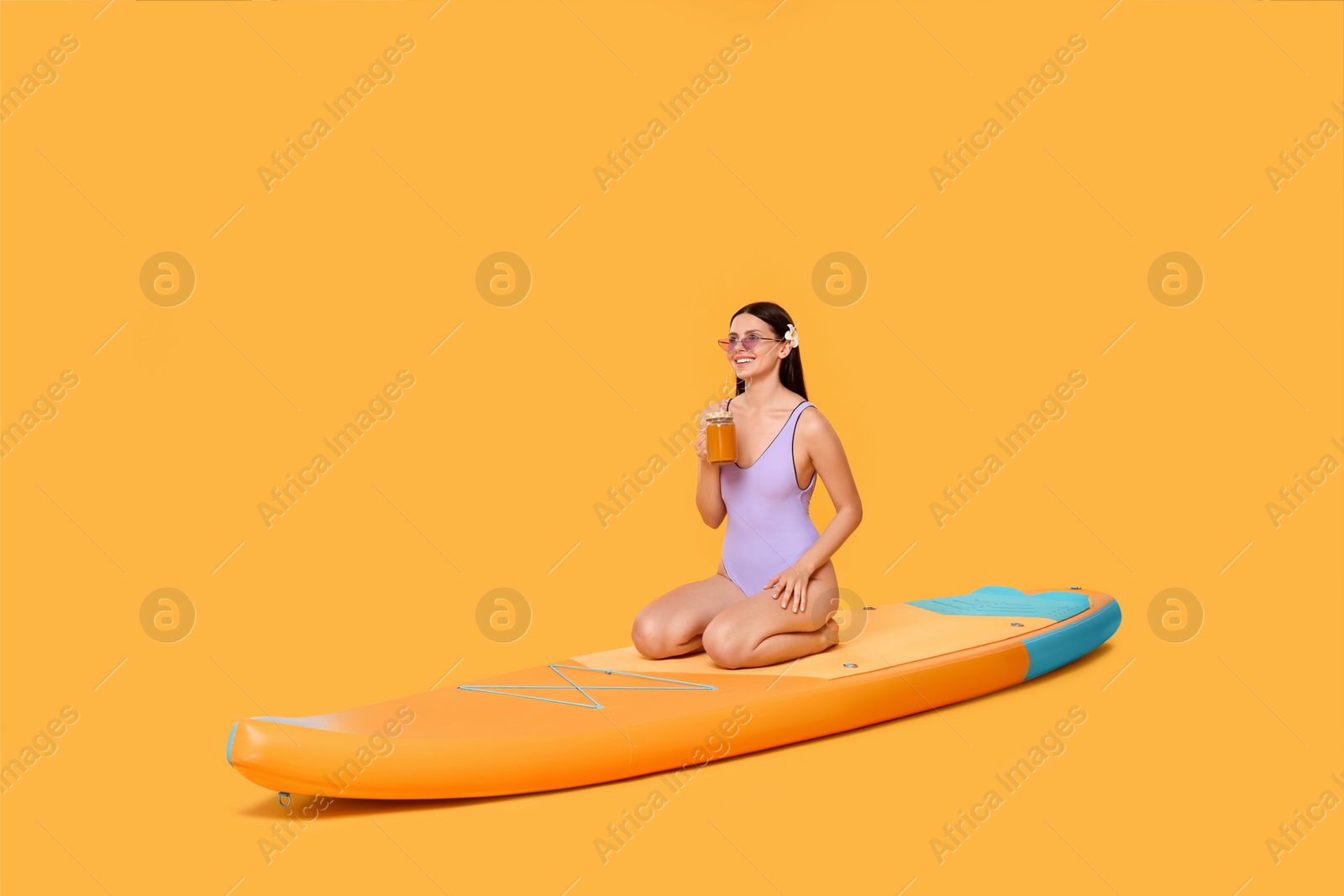 Photo of Happy woman with refreshing drink resting on SUP board against orange background