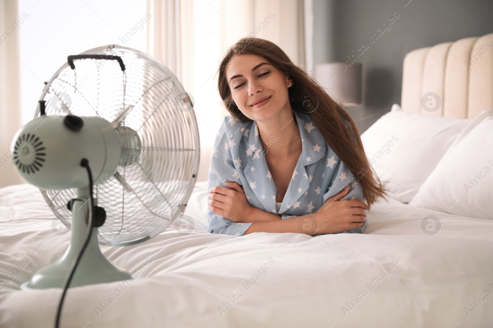 Photo of Woman enjoying air flow from fan on bed in room. Summer heat