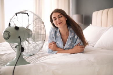 Woman enjoying air flow from fan on bed in room. Summer heat