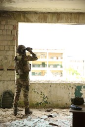 Military mission. Soldier in uniform with binoculars inside abandoned building, back view