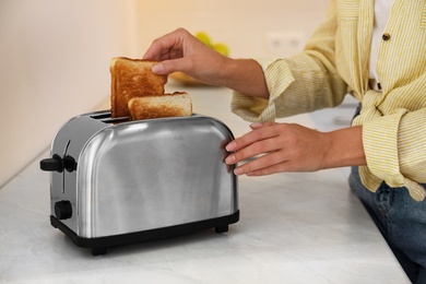 Photo of Woman taking slice of bread from toaster in kitchen, closeup