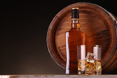 Photo of Whiskey with ice cubes in glass, bottle and barrel on wooden table against dark background, space for text