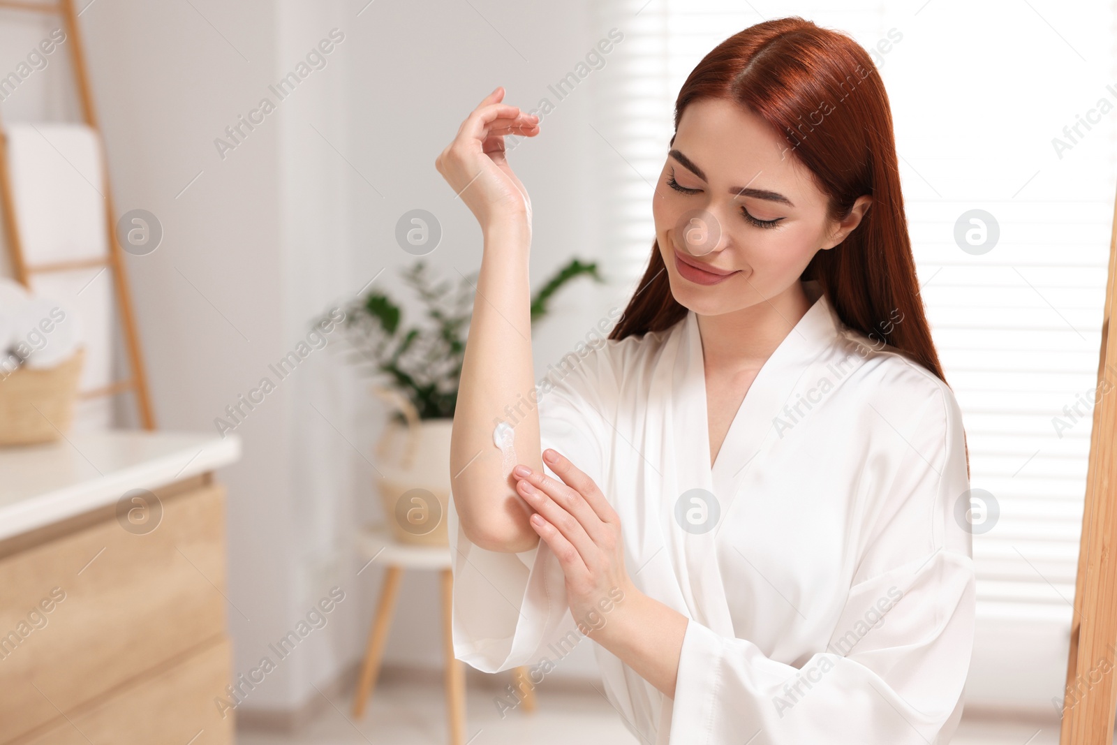 Photo of Beautiful young woman applying body cream onto elbow in bathroom, space for text