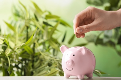 Photo of Woman putting coin into piggy bank on table against blurred background, closeup. Space for text