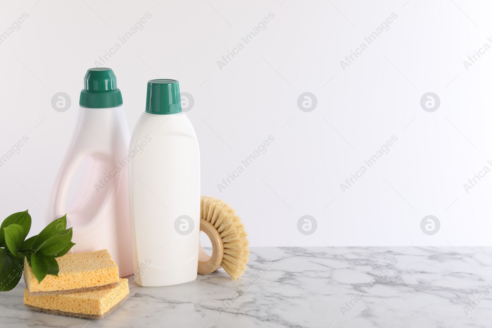 Photo of Bottles of cleaning product, brush, sponges and green leaves on white marble table. Space for text