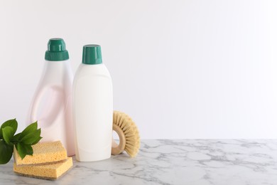 Photo of Bottles of cleaning product, brush, sponges and green leaves on white marble table. Space for text