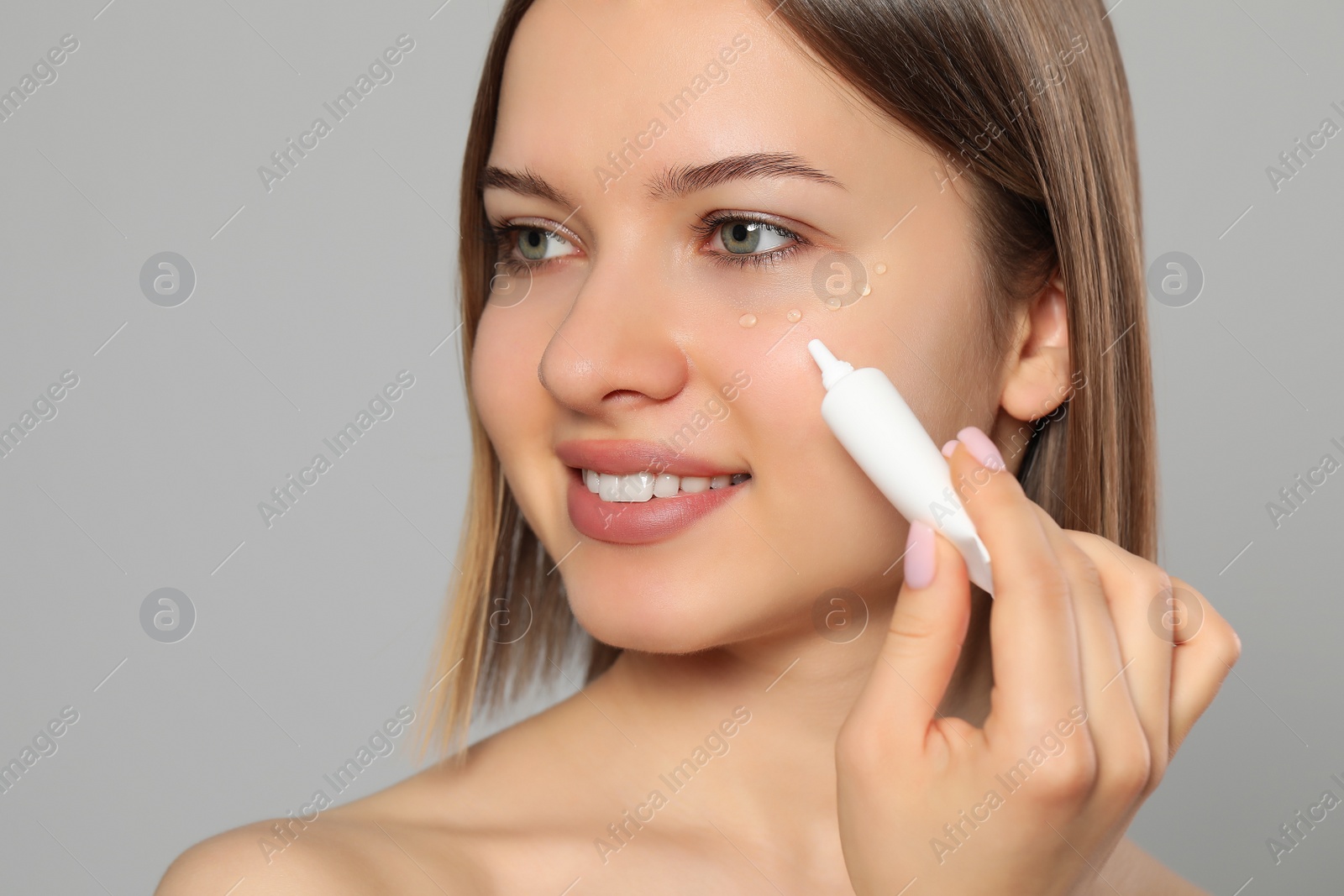 Photo of Young woman applying cream under eyes on light grey background