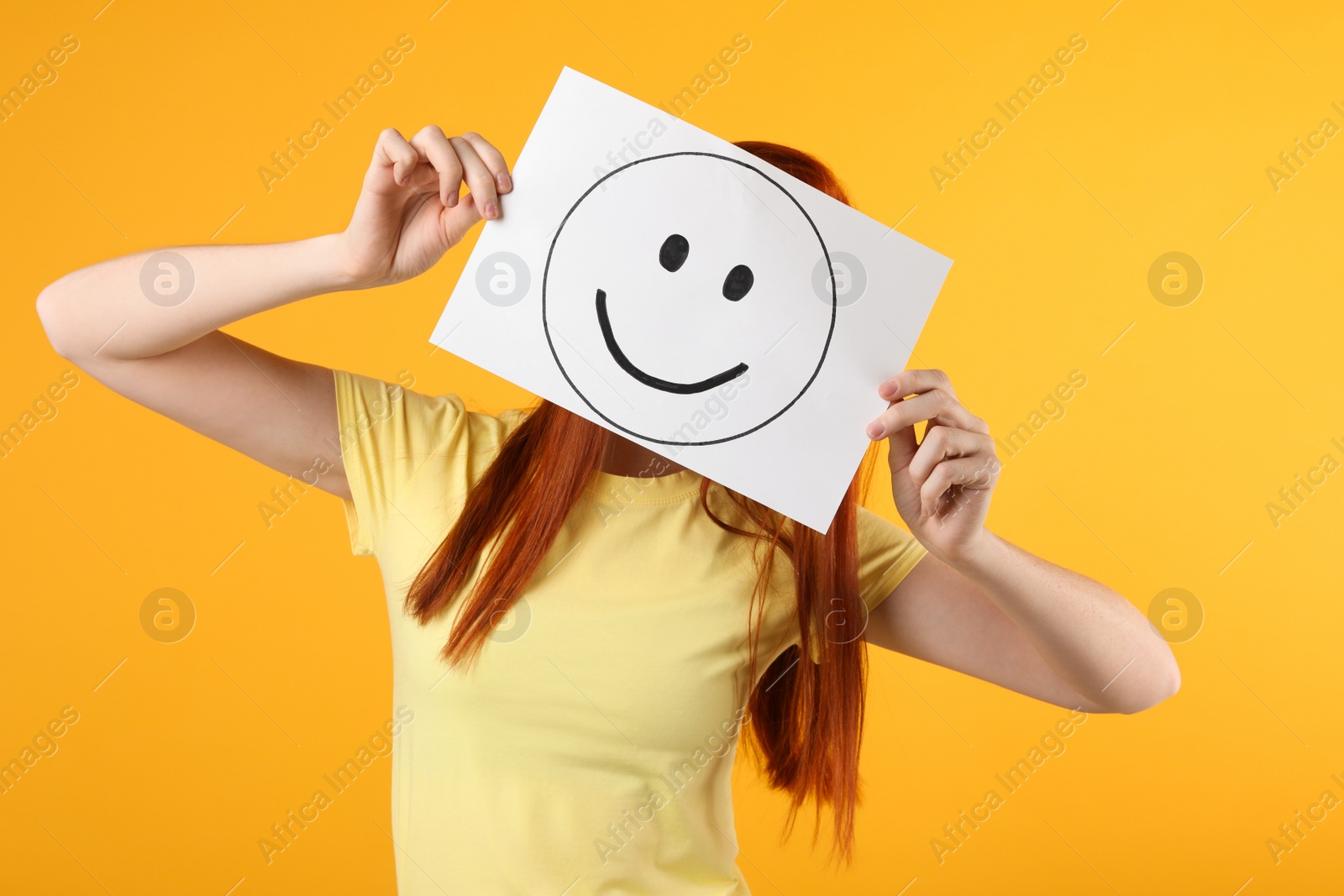 Photo of Woman hiding behind sheet of paper with happy face on yellow background