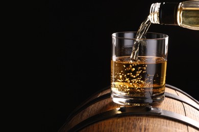 Photo of Pouring whiskey from bottle into glass on wooden barrel against dark background, closeup. Space for text