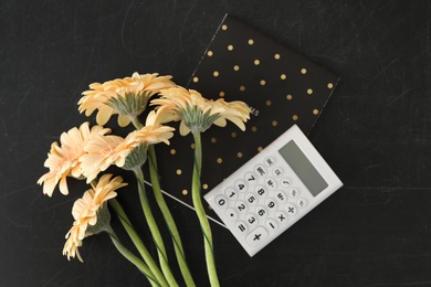 Photo of Notebook, calculator and beautiful flowers on blackboard, flat lay. Happy Teacher's Day