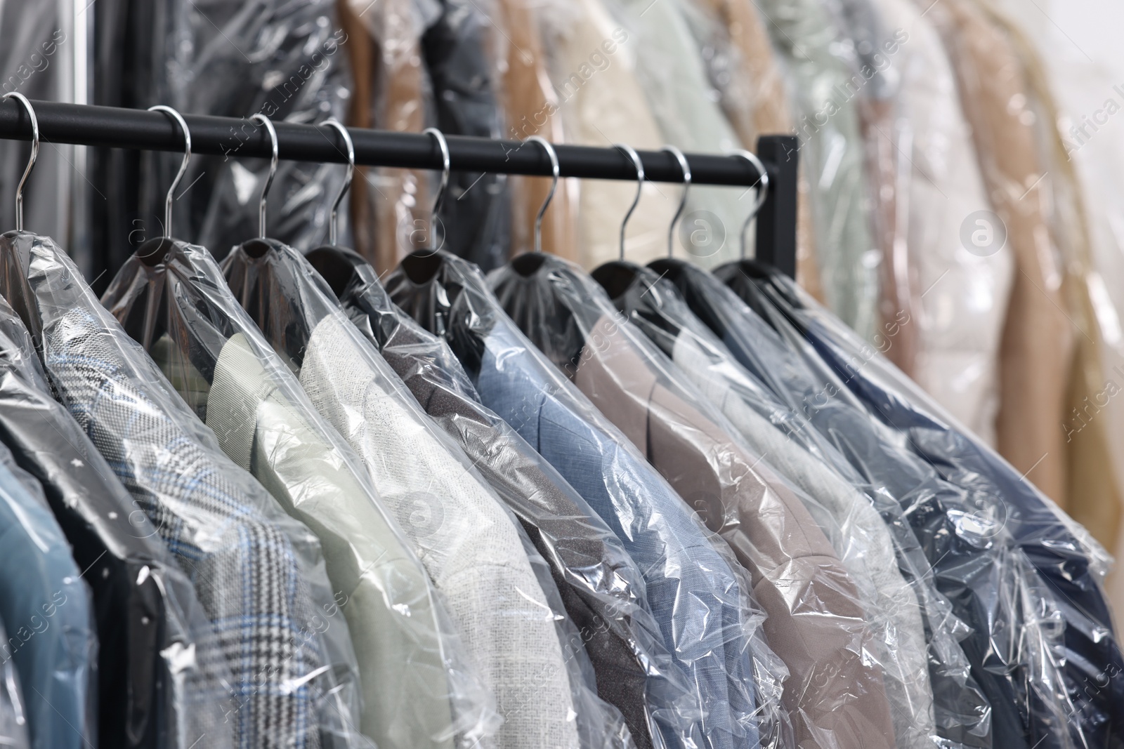 Photo of Dry-cleaning service. Many different clothes in plastic bags hanging on rack indoors, closeup