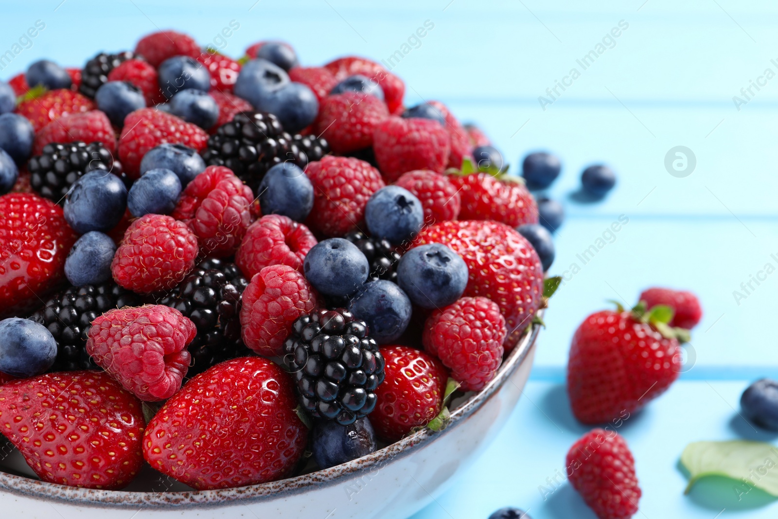 Photo of Different fresh ripe berries in bowl on light blue wooden table, closeup