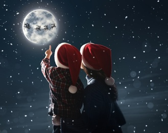 Mother and her little son looking at Santa Claus with reindeers in sky on full moon night. Christmas holiday