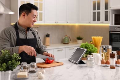 Photo of Cooking process. Man using tablet while cutting fresh bell pepper at countertop in kitchen