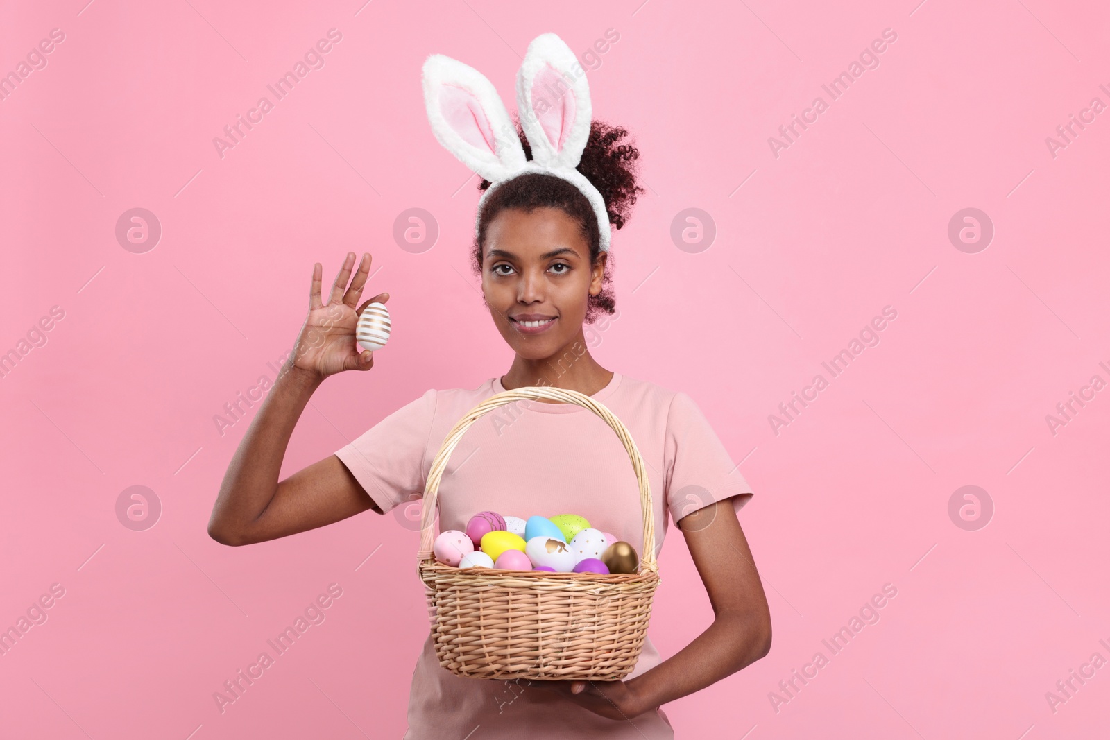 Photo of Happy African American woman in bunny ears headband holding wicker basket with Easter eggs on pink background