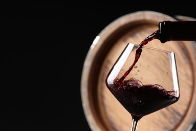 Pouring red wine from bottle into glass near wooden barrel against black background, closeup. Space for text