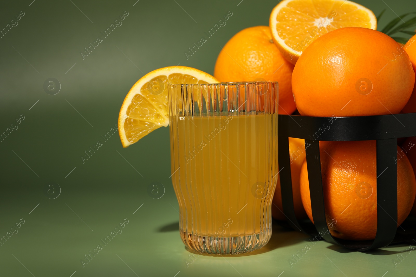 Photo of Fresh oranges in metal basket and glass of juice on green background, closeup