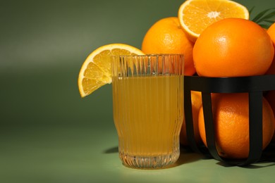 Fresh oranges in metal basket and glass of juice on green background, closeup