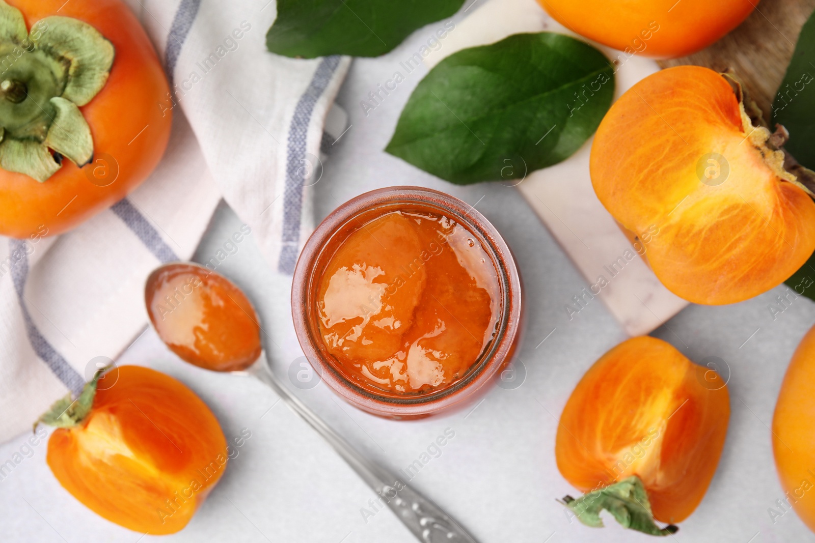 Photo of Jar and spoon of tasty persimmon jam, ingredients on white table, flat lay