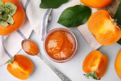 Jar and spoon of tasty persimmon jam, ingredients on white table, flat lay