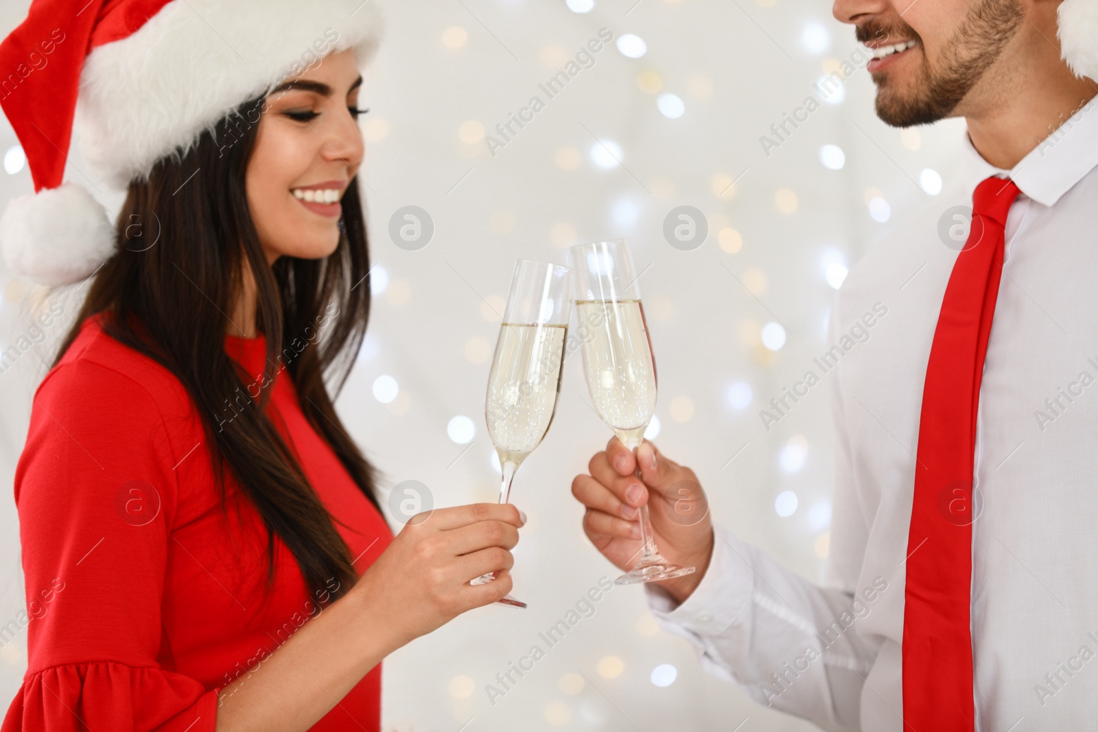 Photo of Happy young couple in Santa hats with glasses of champagne against blurred festive lights. Christmas celebration