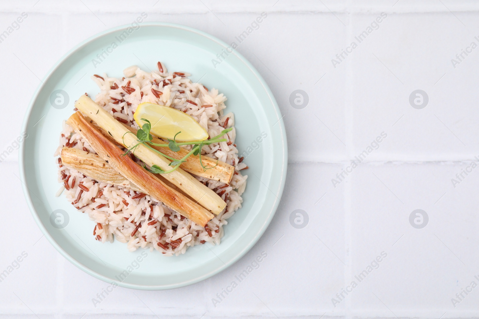 Photo of Plate with baked salsify roots, lemon and rice on white tiled table, top view. Space for text