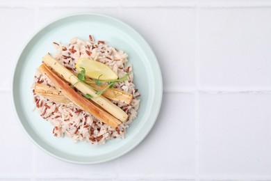 Photo of Plate with baked salsify roots, lemon and rice on white tiled table, top view. Space for text